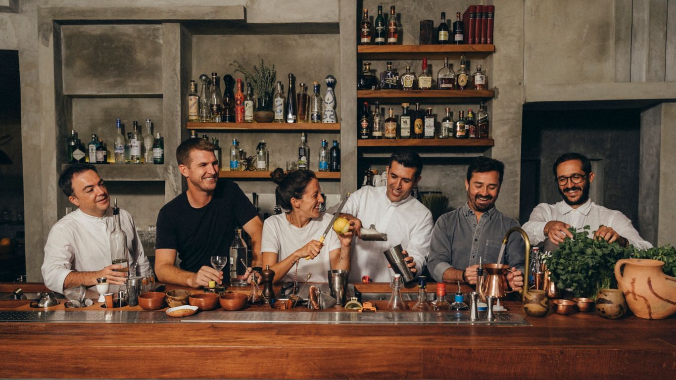 A group of 5 men and 1 woman stand behind a wood bar smiling, laughing and making cocktails